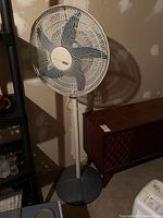 Full view of the Holmes pedestal fan standing with its grey circular base and white pole, set against a wall background.