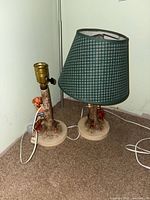 Pair of Goebel Hummel lamps with tree trunk bases and figurines of children, one with a green and white plaid lampshade.