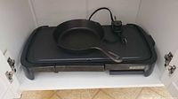 Photo of the black cast iron skillet placed on the griddle surface inside a cabinet, showing the cooking surfaces and handle of the skillet with power cord coiled on the griddle.