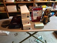 Photo showing assorted candle boxes including taper candles, tealights, and candle holder on ironing board in basement setting.