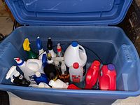 Overview of all bottles and containers inside large blue plastic storage tote showing varied cleaning and pest control chemicals.