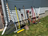 All garden tools lined up outside against siding wall with grass below.