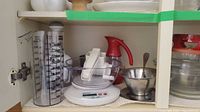 Photo of cabinet shelf with clear plastic measuring cylinders, white digital kitchen scale with bowl, manual white food processor