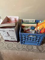 Photo of two boxes containing a mixed assortment of books including children's titles such as 'Caleb's Colt' and 'Cinderella'. Books are arranged vertically and packed in a milk crate and a cardboard box.