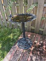Full view of the metal sunflower bird bath with decorative base and small bird figurine on rim, placed outdoors on wooden surface against fence.