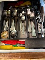 Full view of drawer tray with assorted stainless steel forks, spoons, knives, and plastic handled kitchen utensils including red, yellow handled knives and other colorful utensils.