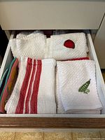 Drawer with folded white dish towels including embroidered tomato and pea pod, a white towel with red stripes, and colorful kitchen linens to the side.