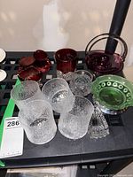 Lot of mixed vintage pressed glassware on black surface including clear textured glasses, green pressed glass plate, and various red glass pieces including a handled basket and cups.