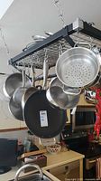 Photo showing hanging cookware including stainless steel pots, pans, strainers on black metal rack.