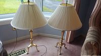 Pair of brass table lamps placed side by side on carpet near window, showing overall appearance, size and matching shades.