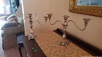 Pair of ornate silver-tone three-arm candelabra on decorative fabric runner on cabinet, showing size and condition.