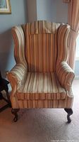 Front view of one wingback armchair showing striped upholstery in beige/cream with multicolor stripes and dark wood legs on carpeted floor near window.