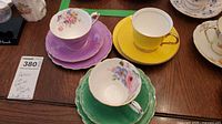 Photo showing three teacup sets with saucers and plates in purple, yellow, and green colors with floral patterns and gold trim.