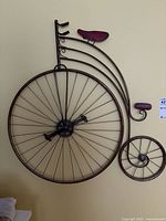 Full view of the metal penny-farthing bicycle wall decor showing the large front wheel with many spokes, smaller rear wheel also with spokes, curved metal frame, seat, and handlebars. Wall mount design with dark brownish bronze finish.
