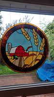 Round stained glass window panel hanging in a window with farm scene including wheat, barn, and tractor in colorful glass pieces outlined in metal leading.