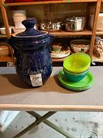 Photo of the dark blue ceramic Rumtopf pot with lid and small stack of green and blue plastic bowls and plates on a table.