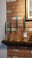 Four brass candlestick holders, two taller and two shorter, each with green taper candles, displayed on a dark wood shelf against a brick wall.