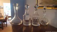 Four crystal decanters arranged in a row on wooden furniture, three have stoppers, one does not