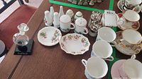 Photo showing salt and pepper shakers, floral and Christmas tree pattern plates, cups, saucers, and silver condiment holders arranged on table.
