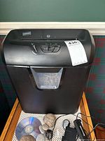 Front view of the black Staples paper shredder showing the transparent window in the bin with shredded paper visible inside, placed on a wooden table with other miscellaneous items around.