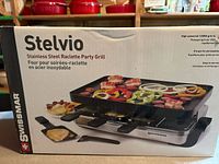 Photo of Swissmar Stelvio Stainless Steel Raclette Party Grill box showing grill with food on top, 8 trays under grill, and spatulas.