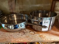 Two stainless steel cookware pieces placed on a wooden shelf, showing a pot and frying pan side by side.
