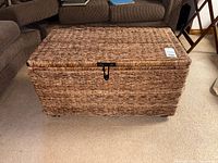 Front and top view of the natural brown wicker/rattan storage trunk chest closed, showing the woven texture and metal clasp.