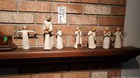 Seven Willow Tree resin figurines displayed in a row on a wooden mantle against a brick wall, showing size range and detail of each figure.