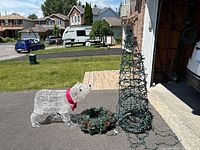 Outdoor setting image showing wire-frame polar bear with red scarf, cone-shaped wire Christmas tree with string lights and star on top, and green wreath with pinecones and red berries.