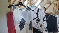 Photo showing multiple women's clothing items on hangers including a white sleeveless dress with floral and abstract black/gray designs, a coral or red long-sleeve top, a black sweater/cardigan with neckline detail, and several garment bags.