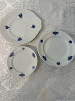 View of one 8.5 inch cake plate, one 7 inch plate, and one 6 inch plate showing the white bone china with blue grape cluster design, placed on a white patterned tablecloth.