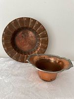 Both antique copper items displayed on white fabric: a small copper pot with scalloped silver rim, and a hammered decorative hanging bowl with scalloped edge.