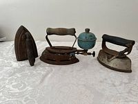 Lot of three antique sad irons on a white tablecloth including a blue fuel tank gas iron and two cast irons
