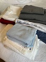 Stack of various folded bedding including white, cream, light blue, grey, and red colored items, some appearing as sheets and pillowcases.