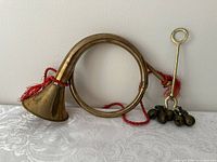 Photo showing coiled brass horn with flared bell and attached red cord alongside seven brass bells on a brass handle with a loop and red cord.