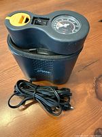 Front view of compact black air compressor showing built-in pressure gauge, on/off switch, and attached cable.