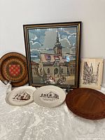 Photo showing the framed needlepoint of St. Mark's Church with distinctive roof tiles, carved Croatian coat of arms plaque, carved wooden plate with French inscription, two church-themed plates, angel figure, small cross, and street scene print.