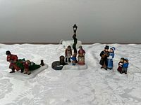 Three porcelain Dickens’ Village figurines displayed on white fabric background: man with Christmas tree and child, family by lamp post with coal barrel, Victorian couples and children