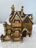 Front view of the Dept 56 Dickens Village Tattyeave Knoll porcelain house illuminated from inside with light cord, snow on roof, and three red figurines standing outside.