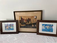 Three framed prints with wooden frames, two smaller and one large, displaying various artistic cityscape and village scenes.