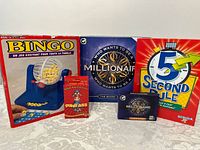 Photo showing all 5 games in lot: Bingo game with cage, Who Wants to Be a Millionaire board game, 5 Second Rule game, Dumb Ass card game, and a Who Wants to Be a Millionaire card game box.