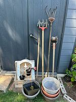 Three long-handled garden tools and a stack of plant pots, with garden decor items visible in front of a dark shed door.