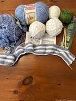 Photo of multiple yarn balls in blue, white, cream, and green, wooden knitting needles, partially knitted blue and white striped baby blanket, and instructions visible beneath.