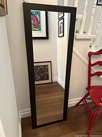 Front view of the full-length rectangular mirror with black wooden frame leaning against wall beside red chair and artwork.