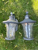Two metal outdoor porch light fixtures laying on grass, both showing signs of fading and wear, with clear textured glass panels.
