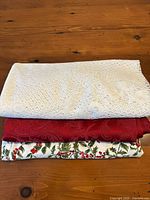 Three folded round tablecloths on a wooden surface showing lace, red holly-embossed, and holly patterned cloths.