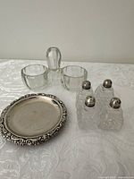 Full view showing four salt and pepper shakers with glass bodies and sterling silver tops, vintage glass salt and pepper holder with toothpick holder at the center, and four silver-plated coasters with ornate edges.