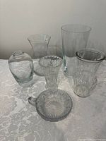 Seven various clear glass and crystal vases of different shapes and a small decorative dish on a table with a white lace cloth