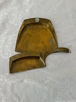 Both brass pieces, dustpan and scraper, shown side by side on white patterned surface.