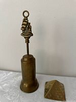 Photo showing the brass fireside brush with closed retractable bristles and pyramid paperweight on a white surface.